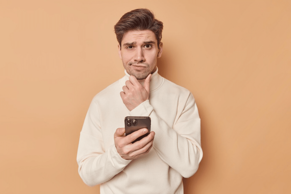 An attractive man wearing a light sweater, holding his chin with one hand, and looking puzzled while holding a phone in the other.