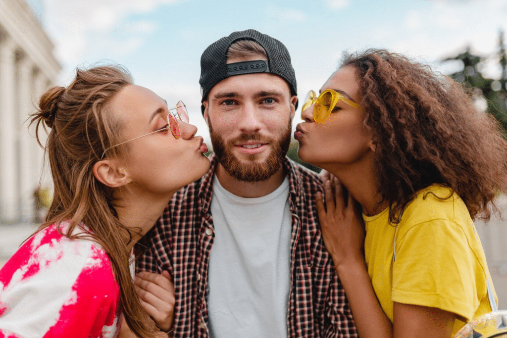 An attractive man wearing a cap, posing with two attractive girls on each side wearing sunglasses, about to kiss him.