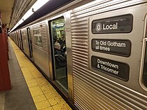 A corrugated silver metal subway train sits with its doors open in a station. Its rollsign reads 0 Local / To Old Gotham all times / Downtown & Tricorner.