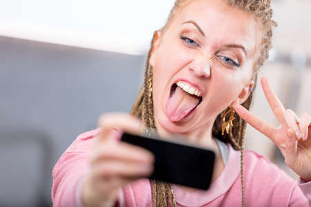 Young woman pulling a goofy face for a selfie sticking out her tongue and making a V-sign gesture as she poses for her mobile phone camera Stock Photo - 103269368
